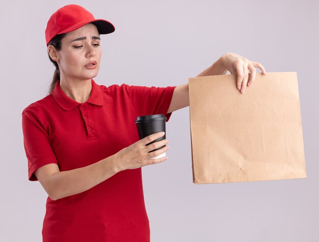 Confusa giovane donna delle consegne in uniforme e cappuccio che tiene una tazza di caffè in plastica e un pacchetto di carta guardando la tazza di caffè isolata sul muro bianco