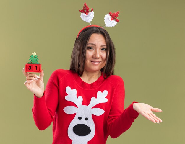 Confusa giovane ragazza asiatica indossando il cerchio dei capelli di Natale che tiene la mano di diffusione del giocattolo di Natale isolata su fondo verde oliva