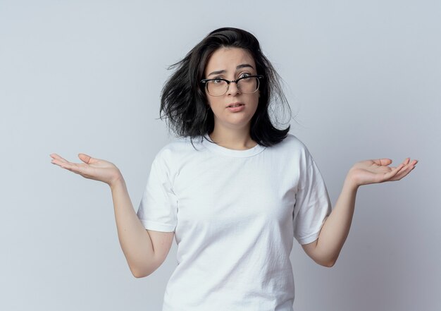 Confusa giovane ragazza piuttosto caucasica con gli occhiali che mostra le mani vuote guardando la telecamera isolata su sfondo bianco