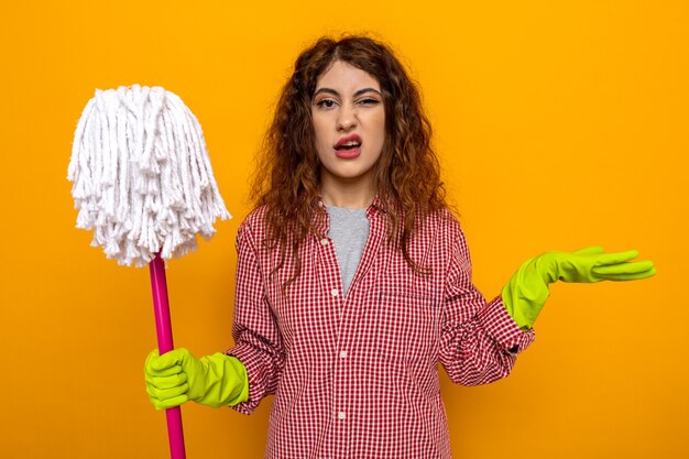 Confusa mano di diffusione giovane donna delle pulizie che indossa guanti che tengono mop isolato su parete arancione