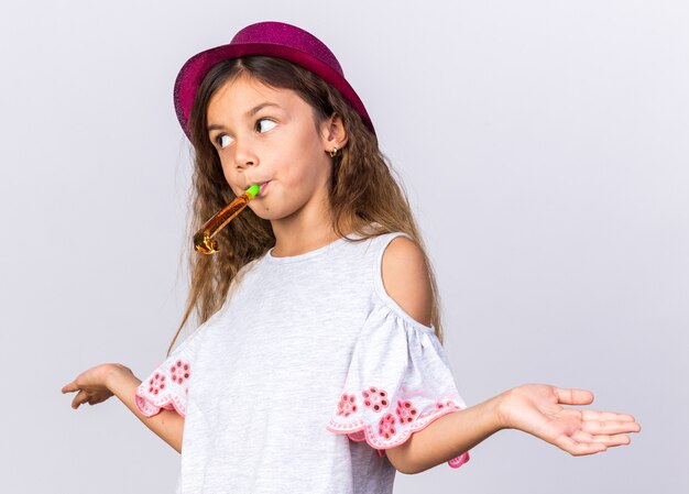 confusa piccola ragazza caucasica con cappello da festa viola che soffia fischio di partito guardando il lato isolato sul muro bianco con lo spazio della copia