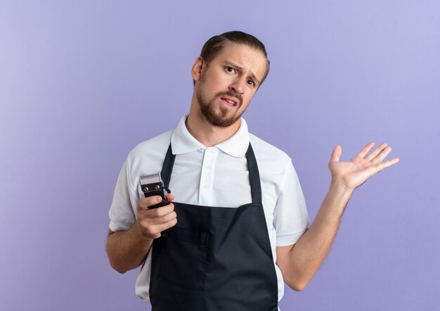 Confuso giovane barbiere bello che indossa uniformi tenendo i tagliacapelli e mostrando la mano vuota isolata sulla porpora con lo spazio della copia