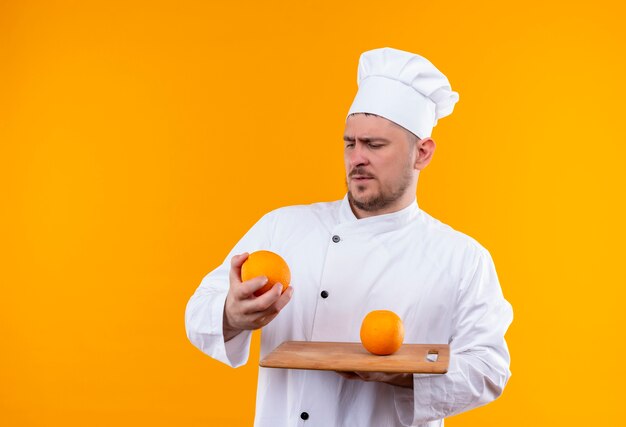 Confuso giovane bel cuoco in uniforme da chef che tiene tagliere e arancia e lo guarda isolato su una parete arancione orange
