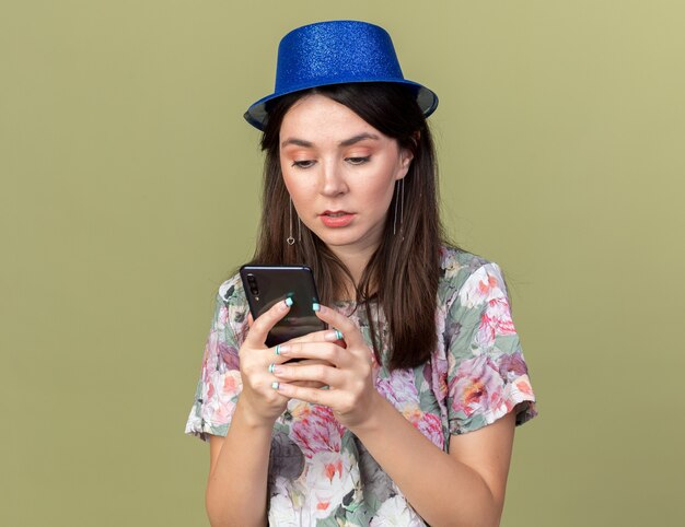 Confuso giovane bella donna che indossa cappello da festa tenendo e guardando il telefono isolato su parete verde oliva