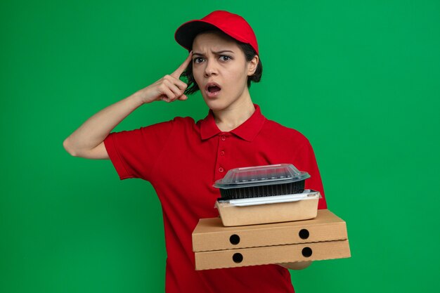 Confuso giovane bella donna di consegna che tiene contenitori per alimenti e imballaggi su scatole per pizza