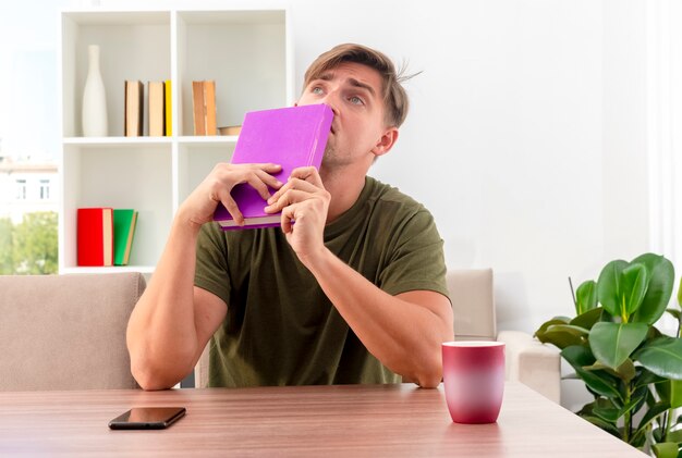 Confuso giovane biondo bell'uomo si siede al tavolo con il telefono e la tazza che tiene il libro vicino alla bocca e alzando lo sguardo all'interno del soggiorno