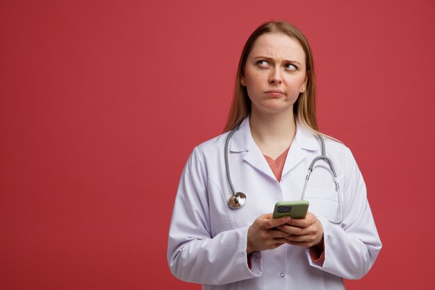 Confuso giovane dottoressa bionda che indossa veste medica e stetoscopio intorno al collo utilizzando il telefono cellulare cercando con le labbra increspate