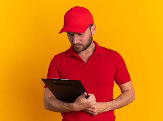 Confuso giovane fattorino caucasico in uniforme rossa e berretto che tiene e guarda la lavagna per appunti isolata sulla parete arancione