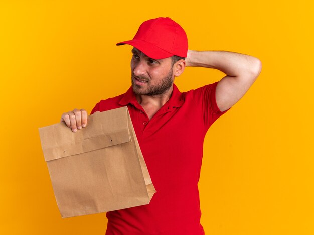 Confuso giovane fattorino caucasico in uniforme rossa e cappuccio che tiene il pacchetto di carta tenendo la mano dietro la testa guardando il lato isolato sul muro arancione