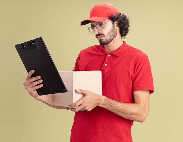 Confuso giovane fattorino in uniforme rossa e berretto con gli occhiali che tengono scatola di cartone e appunti guardando gli appunti isolati sul muro verde oliva