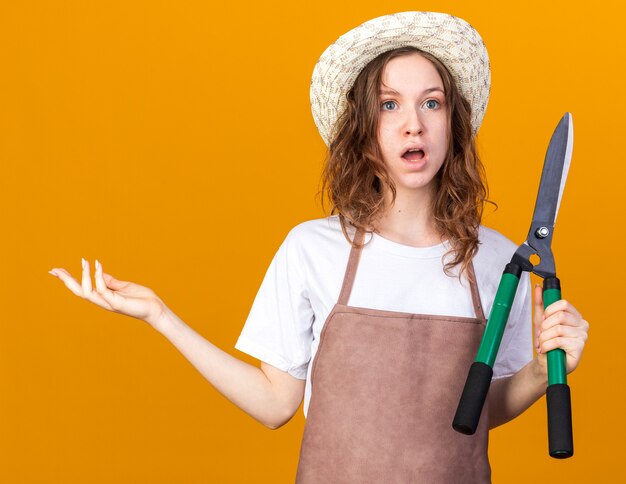 Confuso giovane giardiniere femminile che indossa cappello da giardinaggio che tiene la mano di diffusione del taglio di potatura isolata sulla parete arancione