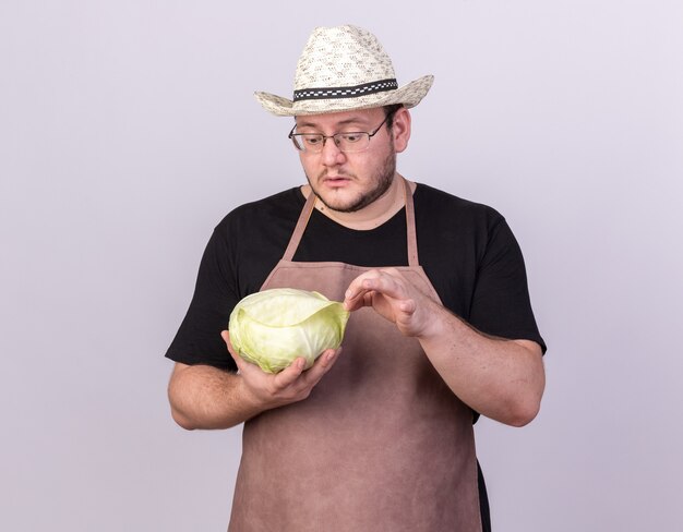 Confuso giovane giardiniere maschio che indossa cappello da giardinaggio tenendo e guardando cavolo isolato su muro bianco