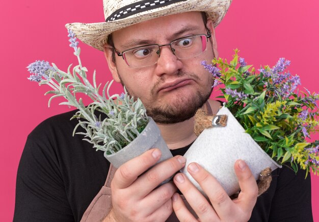 Confuso giovane giardiniere maschio che indossa cappello da giardinaggio tenendo e guardando i fiori in vasi da fiori isolati su parete rosa