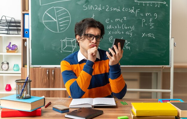 confuso giovane insegnante di geometria con gli occhiali seduto alla scrivania con materiale scolastico in classe tenendo la mano sul mento tenendo e guardando la calcolatrice