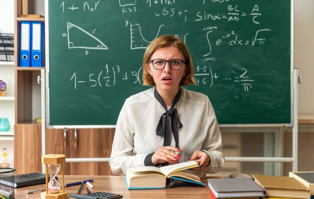 Confuso giovane insegnante femminile si siede al tavolo con materiale scolastico in classe