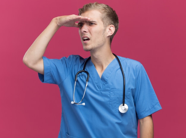 Confuso giovane medico maschio che indossa l'uniforme del medico con lo stetoscopio guardando a distanza con la mano isolata sulla parete rosa