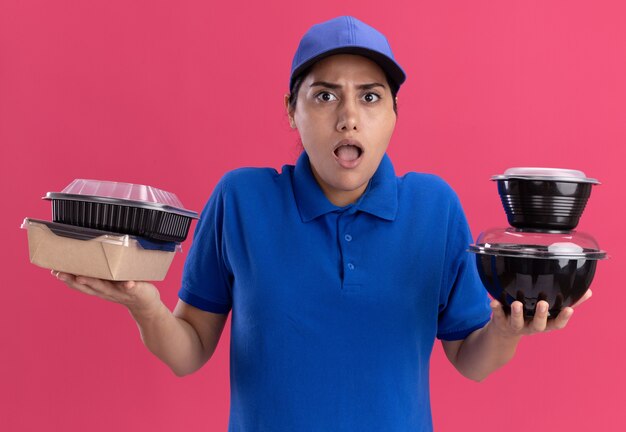 Confuso giovane ragazza delle consegne che indossa l'uniforme con il cappuccio che tiene i contenitori per alimenti isolati sulla parete rosa