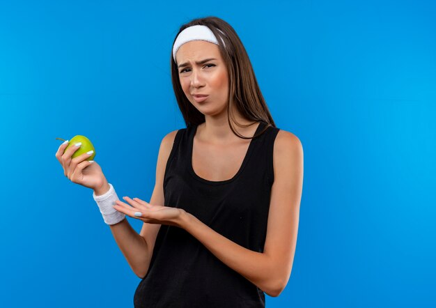 Confuso giovane ragazza piuttosto sportiva che indossa fascia e cinturino che tiene e indica la mela isolata sulla parete blu con spazio copia