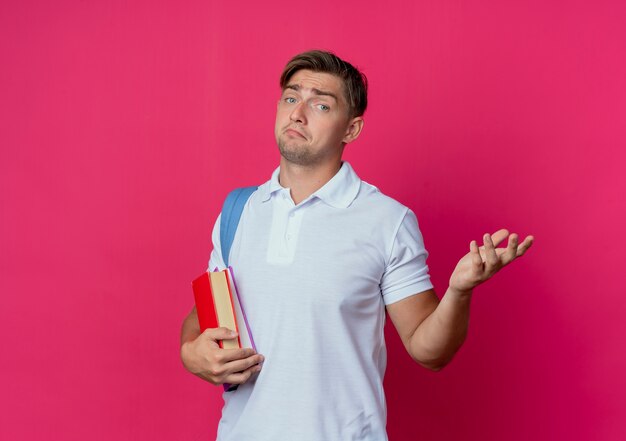 Confuso giovane studente maschio bello che indossa la borsa posteriore che tiene i libri e diffonde la mano isolata sulla parete rosa