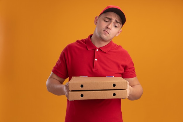 Confuso giovane uomo di consegna che indossa l'uniforme con cappuccio che tiene e guardando le scatole per pizza isolate sulla parete arancione