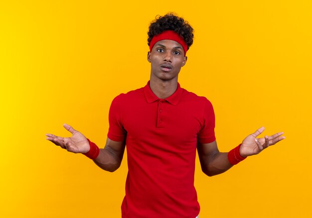 Confuso giovane uomo sportivo afro-americano che indossa la fascia e il braccialetto diffonde le mani isolate su sfondo giallo