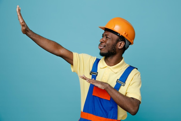 Confuso guardando il lato alzato le mani giovane costruttore afroamericano in uniforme isolato su sfondo blu