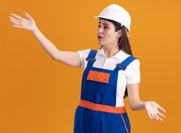 Confuso guardando lato giovane costruttore donna in uniforme diffondendo le mani isolate sulla parete arancione