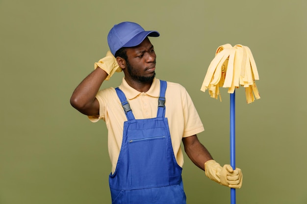 confuso tenendo mop giovane pulitore afroamericano maschio in uniforme con guanti isolati su sfondo verde