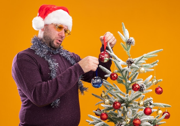 Confuso uomo di mezza età che indossa un cappello da Babbo Natale e una ghirlanda di orpelli intorno al collo con gli occhiali in piedi in vista di profilo vicino all'albero di Natale decorandolo con palline di Natale isolato su sfondo arancione