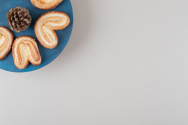 Cono di pino circondato da biscotti a fiocchi su un piatto su sfondo di marmo.