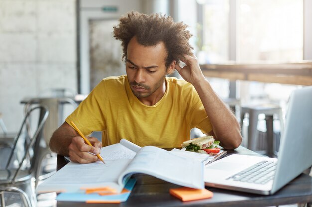 Conoscenza, apprendimento, istruzione e tecnologia. studente con acconciatura afro che risolve problemi matematici, seduto al tavolino del bar con libri di testo e dispositivo elettronico