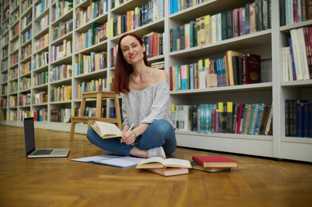 Conoscenza. Giovane donna dai capelli lunghi sorridente che si siede sul pavimento di parquet in biblioteca con il computer portatile e libri che scrivono nello studio del quaderno