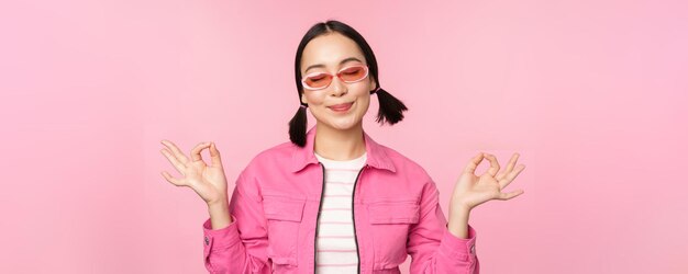 Consapevolezza e concetto di benessere Ragazza coreana sorridente in abito elegante meditando ascoltando mantra tiene le mani in posa di pace zen pratica yoga in piedi su sfondo rosa