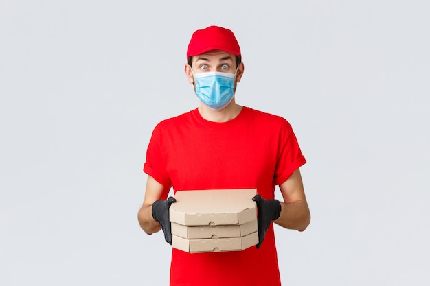 Consegna di cibo, applicazione, drogheria online, shopping senza contatto e concetto di covid-19. Corriere sorpreso in uniforme rossa, maschera facciale e guanti, sembra impressionato, porta la pizza ai clienti, tiene le scatole