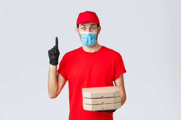 Consegna di cibo, applicazione, drogheria online, shopping senza contatto e concetto di covid-19. Eccitato ragazzo delle consegne in uniforme rossa, guanti e maschera facciale, avere suggerimenti, tenere la pizza e alzare il dito