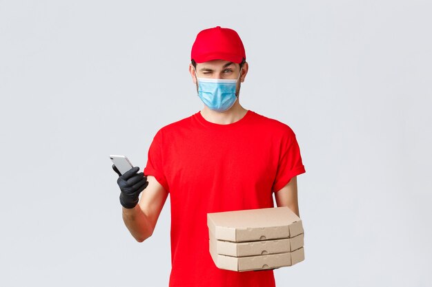 Consegna di cibo, applicazione, shopping senza contatto online e concetto di covid-19. Corriere in divisa rossa, maschera facciale e guanti, strizzando l'occhio al cliente, informare i bonus, sconti speciali sulla pizza, tenere il telefono