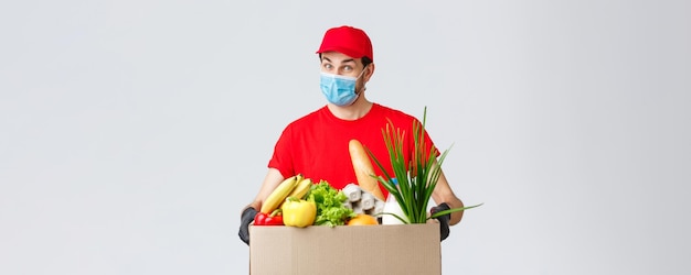 Consegna di generi alimentari e pacchi covid19 quarantena e concetto di shopping Corriere amichevole con maschera facciale e guanti uniforme rossa portare la scatola di cibo al cliente ordinato online consegna senza contatto