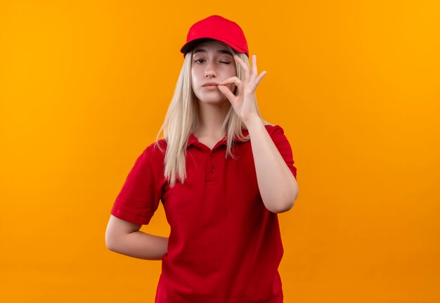 consegna giovane donna che indossa t-shirt rossa e berretto che mostra delizioso gesto sulla parete arancione isolata