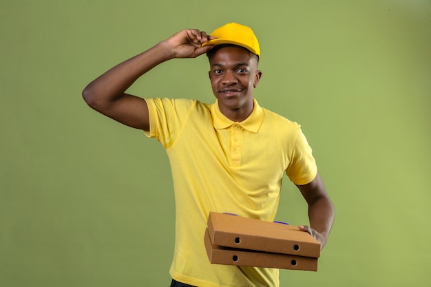 Consegna uomo afroamericano in maglietta polo gialla e berretto in possesso di scatole per pizza saluto toccando il suo berretto sorridente in piedi amichevole sul verde