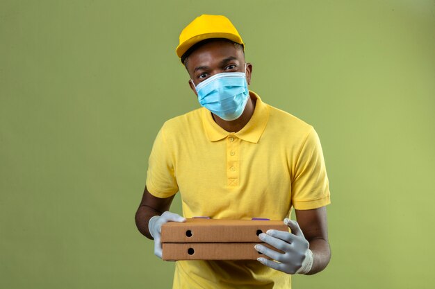 Consegna uomo afroamericano in maglietta polo gialla e berretto indossando maschera protettiva medica che tiene scatole per pizza con il sorriso sul viso in piedi sul verde