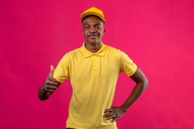 Consegna uomo afroamericano in maglietta polo gialla e berretto sorridente amichevole che mostra i pollici in su sul rosa