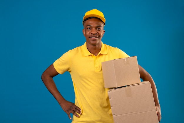 Consegna uomo afroamericano in maglietta polo gialla e berretto tenendo le scatole di cartone con un sorriso amichevole in piedi sul blu isolato