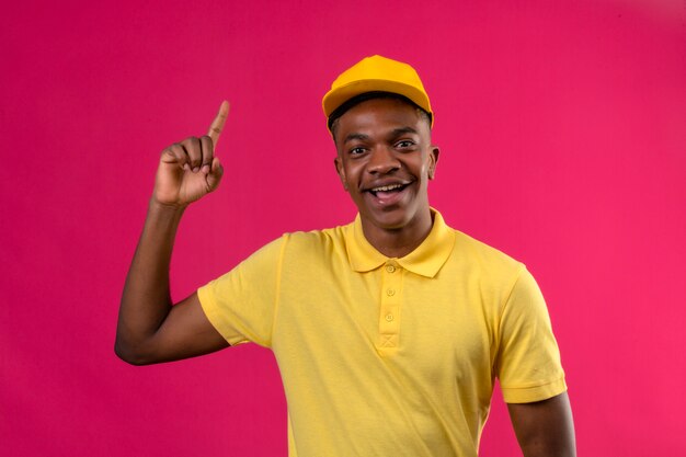 Consegna uomo afroamericano in maglietta polo gialla e cappuccio puntare il dito verso l'alto avendo nuova grande idea sorridendo allegramente sul rosa isolato