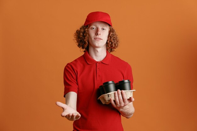 Consegna uomo dipendente in berretto rosso maglietta vuota uniforme tenendo le tazze di caffè guardando la fotocamera sorridente fiducioso allungando la mano in attesa di pagamento in piedi su sfondo arancione
