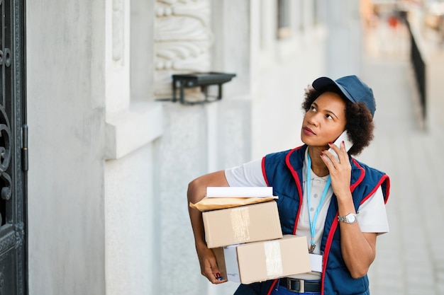 Consegnatore femminile afroamericano che trasporta pacchi e parla al telefono cellulare in città