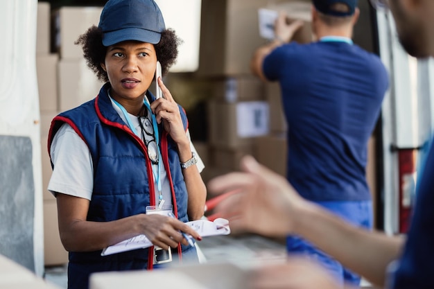 Consegnatore femminile nero che comunica con il collega mentre parla al telefono cellulare e organizza la consegna del pacco