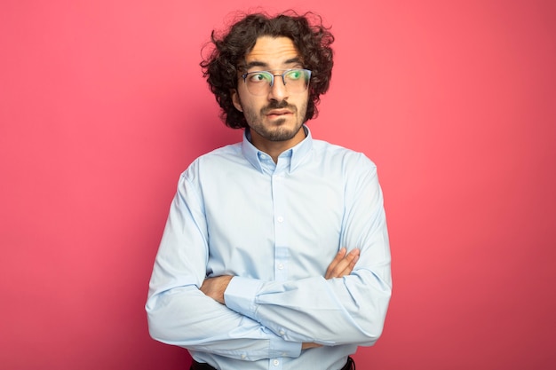 Considerato giovane uomo caucasico bello con gli occhiali in piedi con la postura chiusa guardando il labbro mordace laterale isolato su sfondo cremisi con spazio di copia