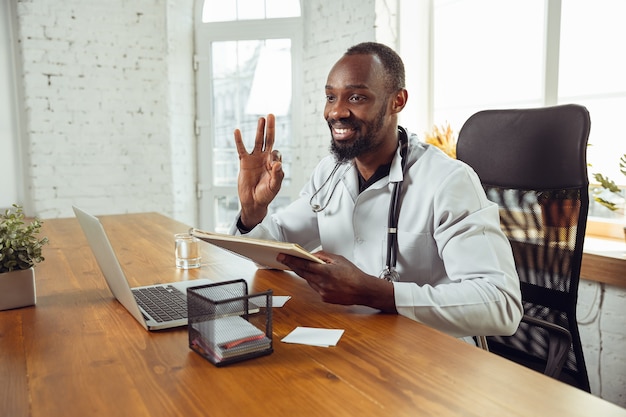 Consulenza medica per il paziente, raccomandazione. Medico afroamericano durante il suo lavoro con i pazienti, spiegando le ricette per la droga. Duro lavoro quotidiano per la salute e il salvataggio di vite durante l'epidemia.