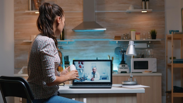 Consultazione di telemedicina durante la pandemia di covid di notte, donna seduta davanti al laptop in cucina. Signora malata che discute durante la consultazione virtuale sui sintomi che tengono una bottiglia di pillole
