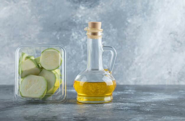 Contenitore di plastica pieno di pezzi di zucchine e bottiglia di olio su sfondo grigio.